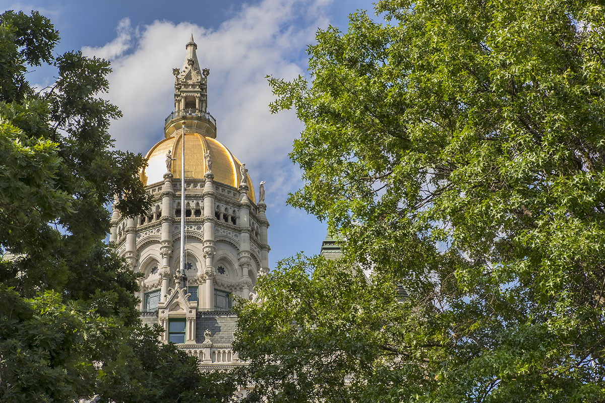 Connecticut State Capitol dome, with Gothic architecture and gold accents, seen from a distance through a gap in bright green tree foliage